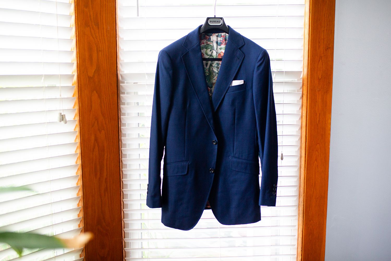 A navy blue blazer with notched lapels hangs against a wall with white louvered shutters in sunlight.