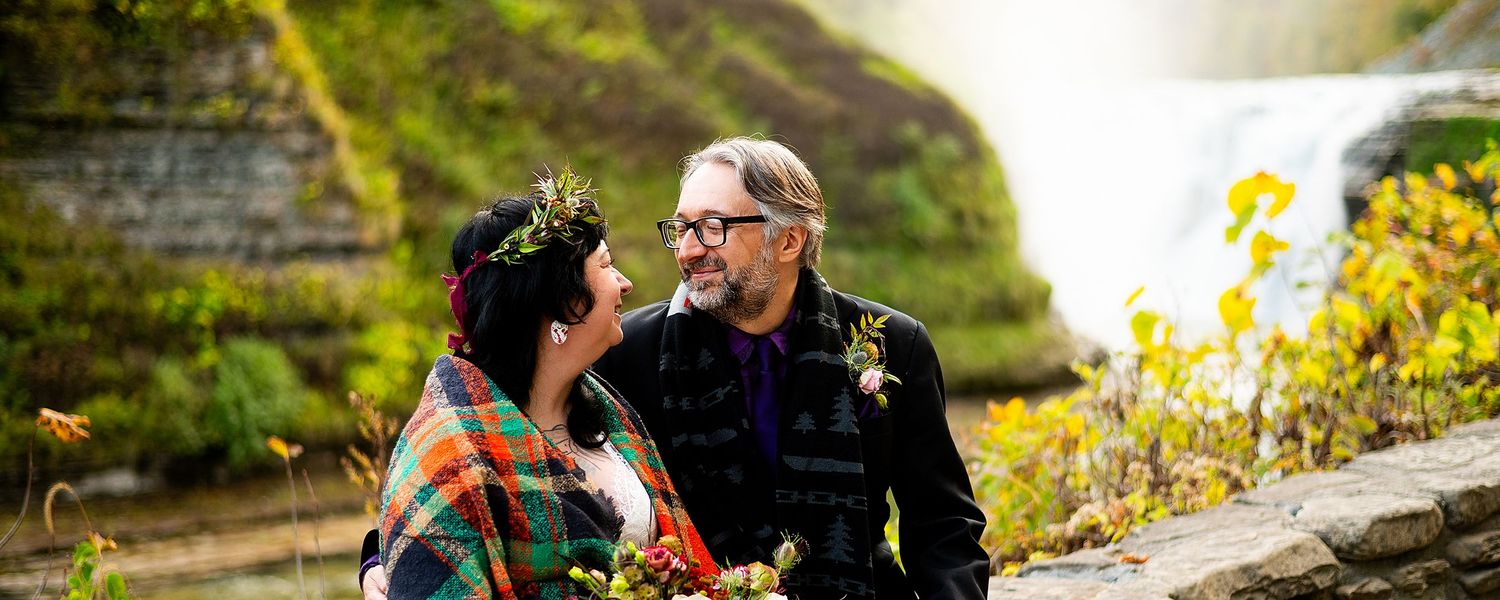A couple shares a romantic moment near a waterfall surrounded by vibrant yellow flowers and lush greenery.