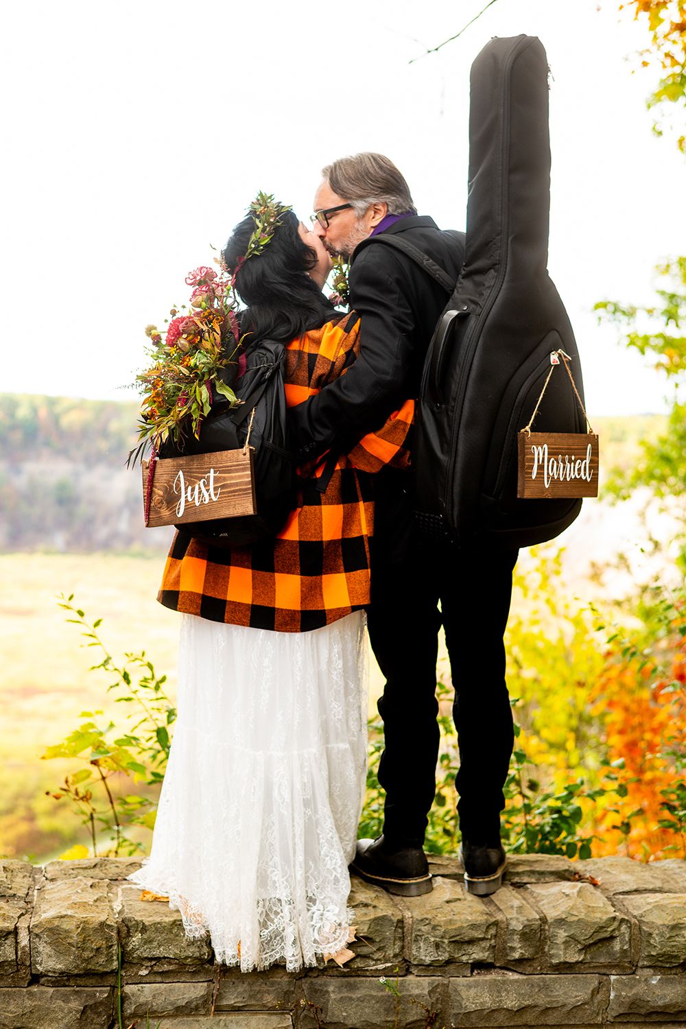 A couple in matching buffalo plaid and black attire embrace on a stone wall with fall foliage and a guitar case in background.