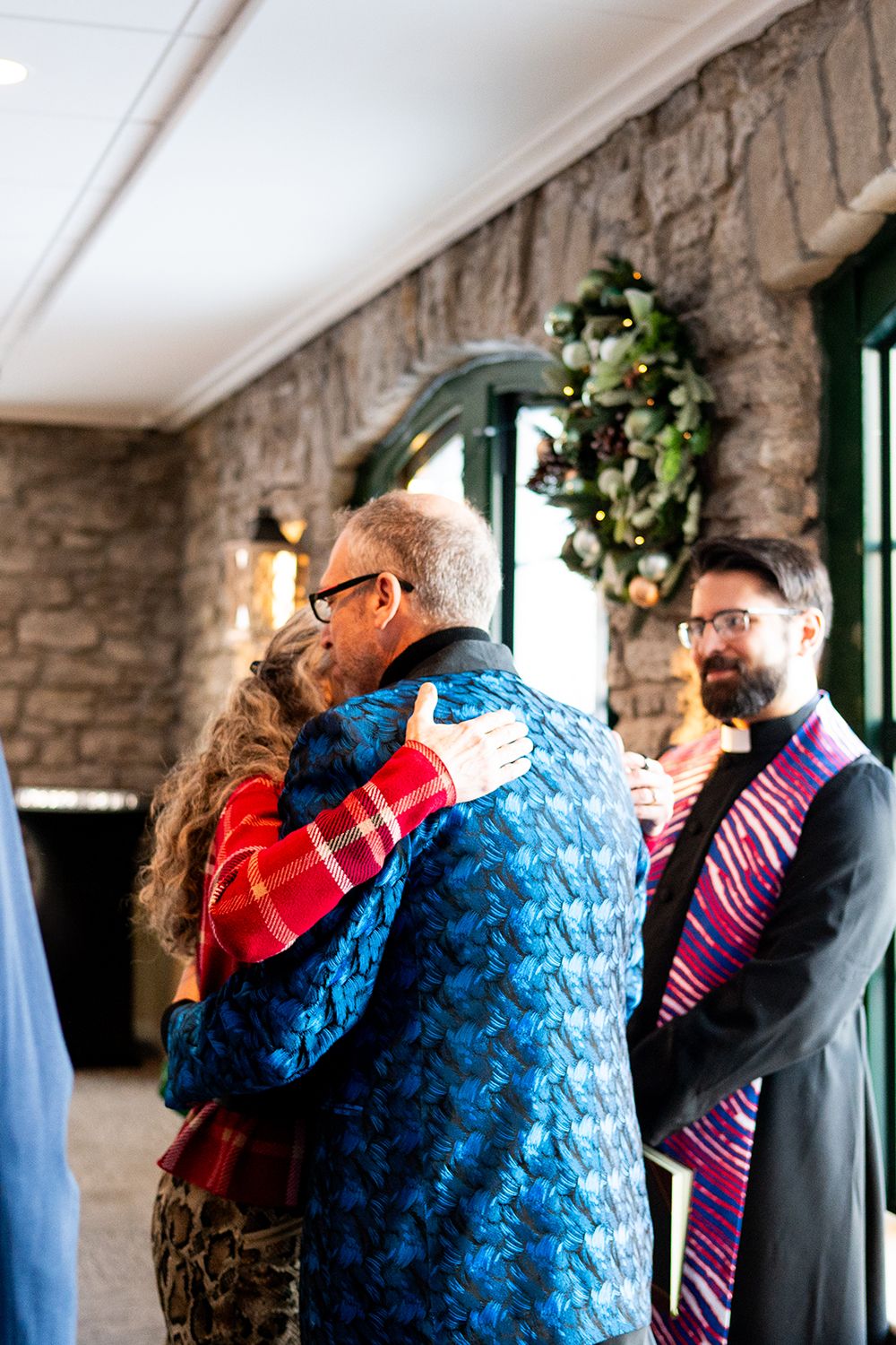 People embrace during an indoor celebration at a stone-walled venue decorated with greenery and warm lighting.