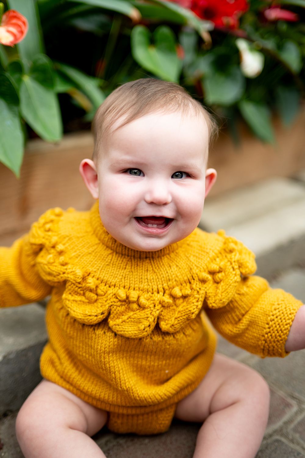 Baby in bright yellow ruffled top sits on stone paving outdoors.
