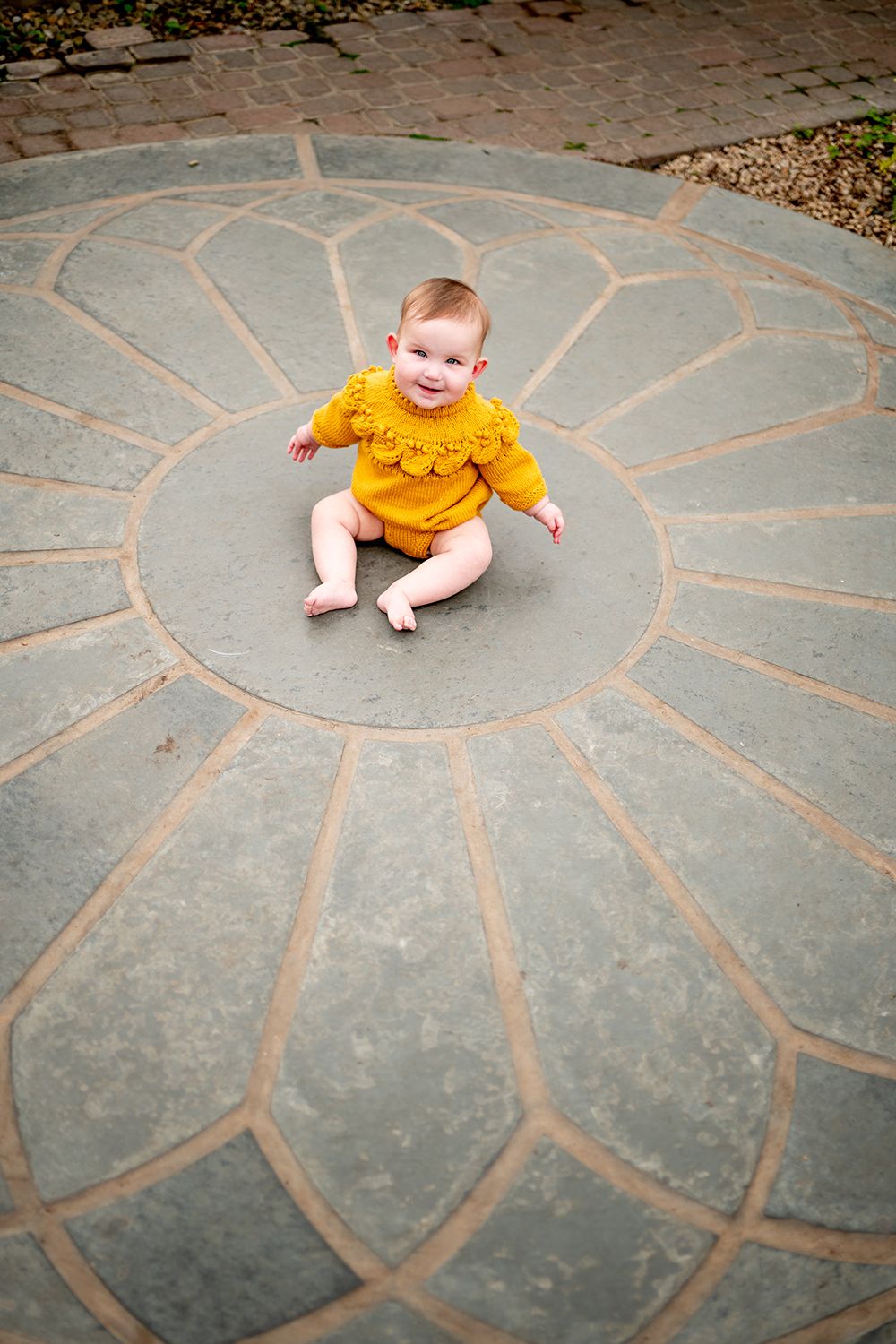 A chalk drawing of a sun design on concrete pavement with a small child in a yellow shirt sitting next to it.