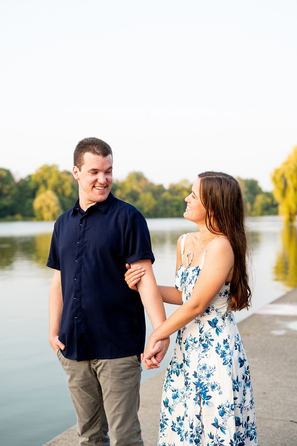 A couple shares a playful moment by a scenic lake with willow trees during golden hour.