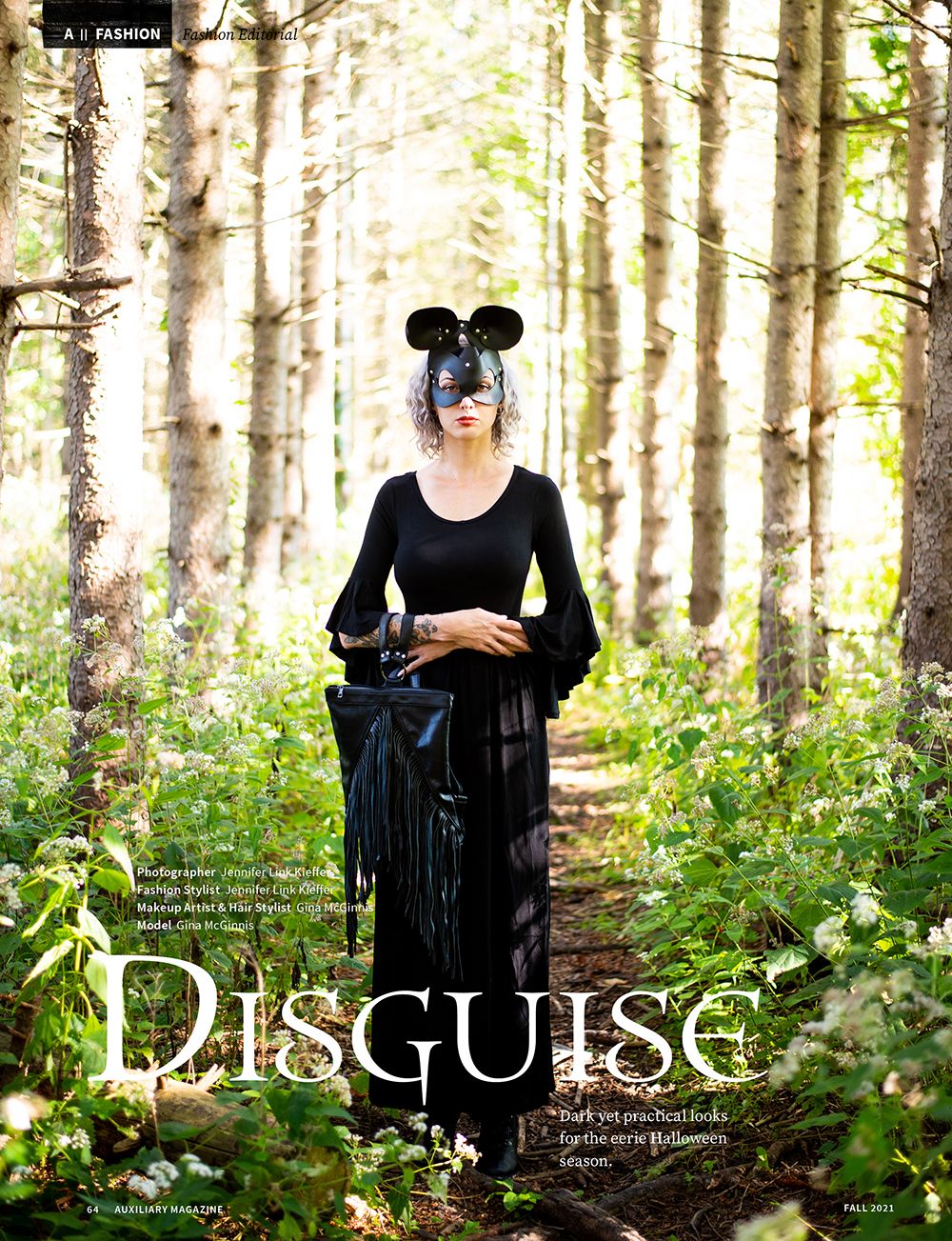 Figure in black outfit with mouse ears and mask stands on forest path surrounded by white birch trees and wildflowers.