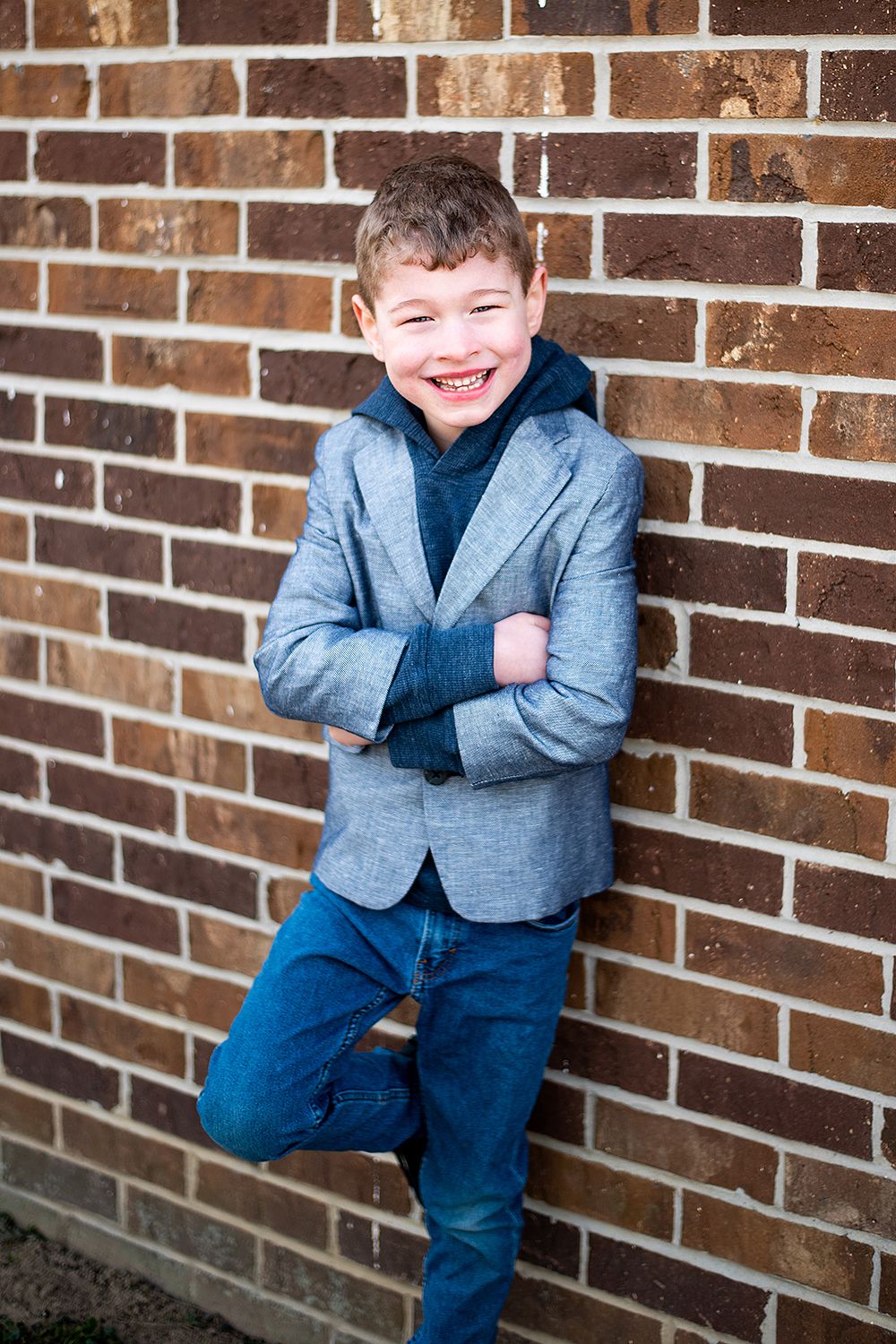 A young figure in a gray blazer and jeans leans casually against a brick wall while smiling warmly.