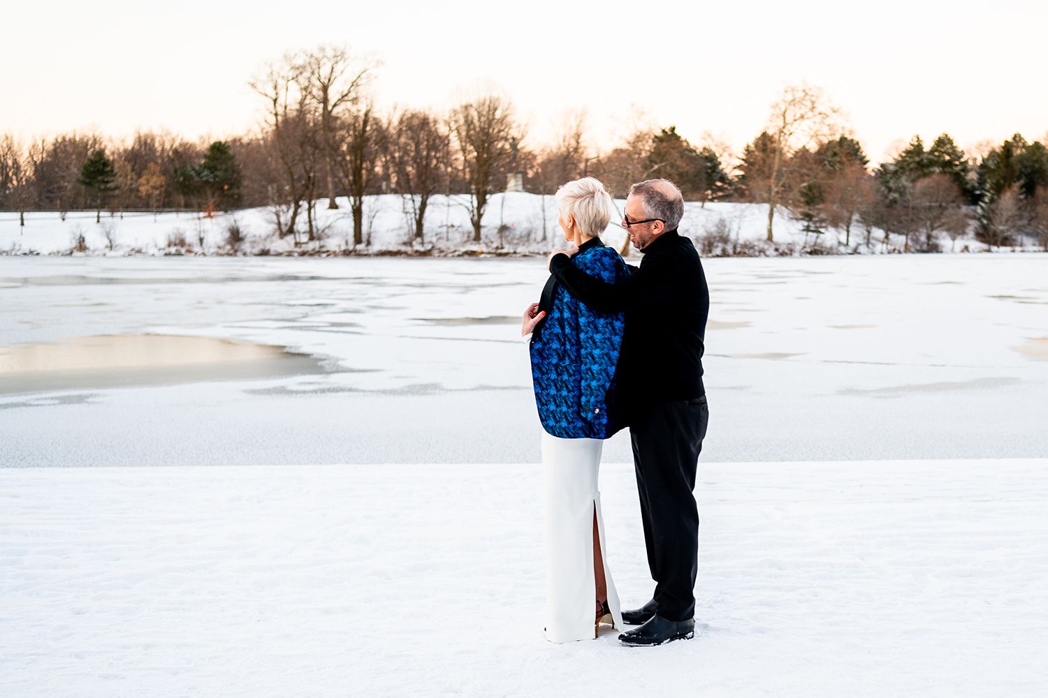 Couple embraces on a snowy winter landscape at sunset near a frozen lake.