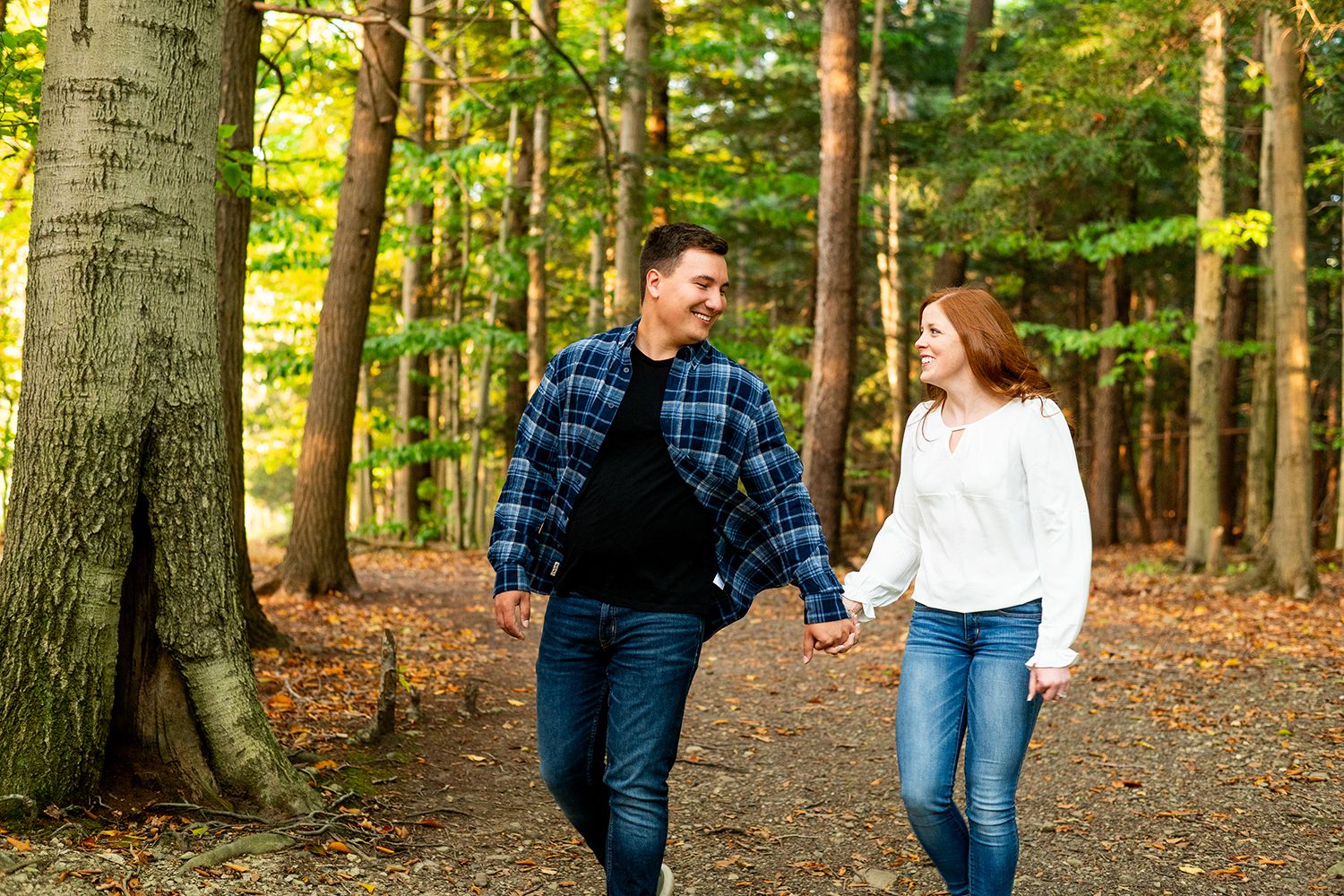 A couple walks hand in hand through a wooded autumn forest path with fallen leaves on the ground.