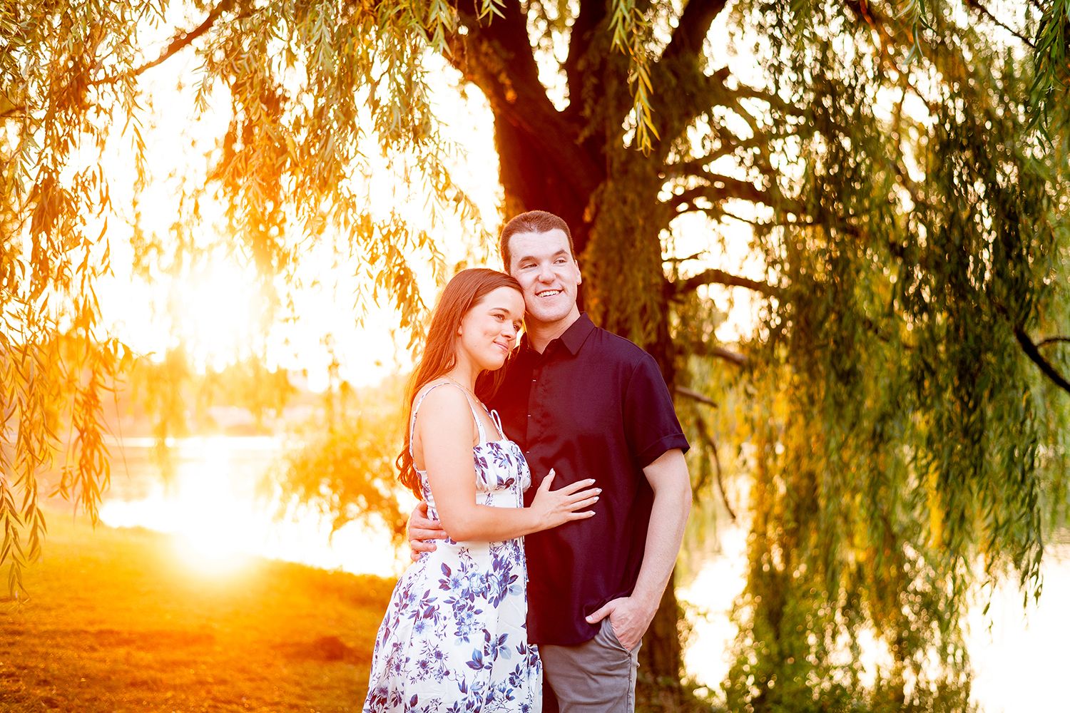A couple embraces outdoors at sunset beneath a weeping willow tree creating a warm golden atmosphere.