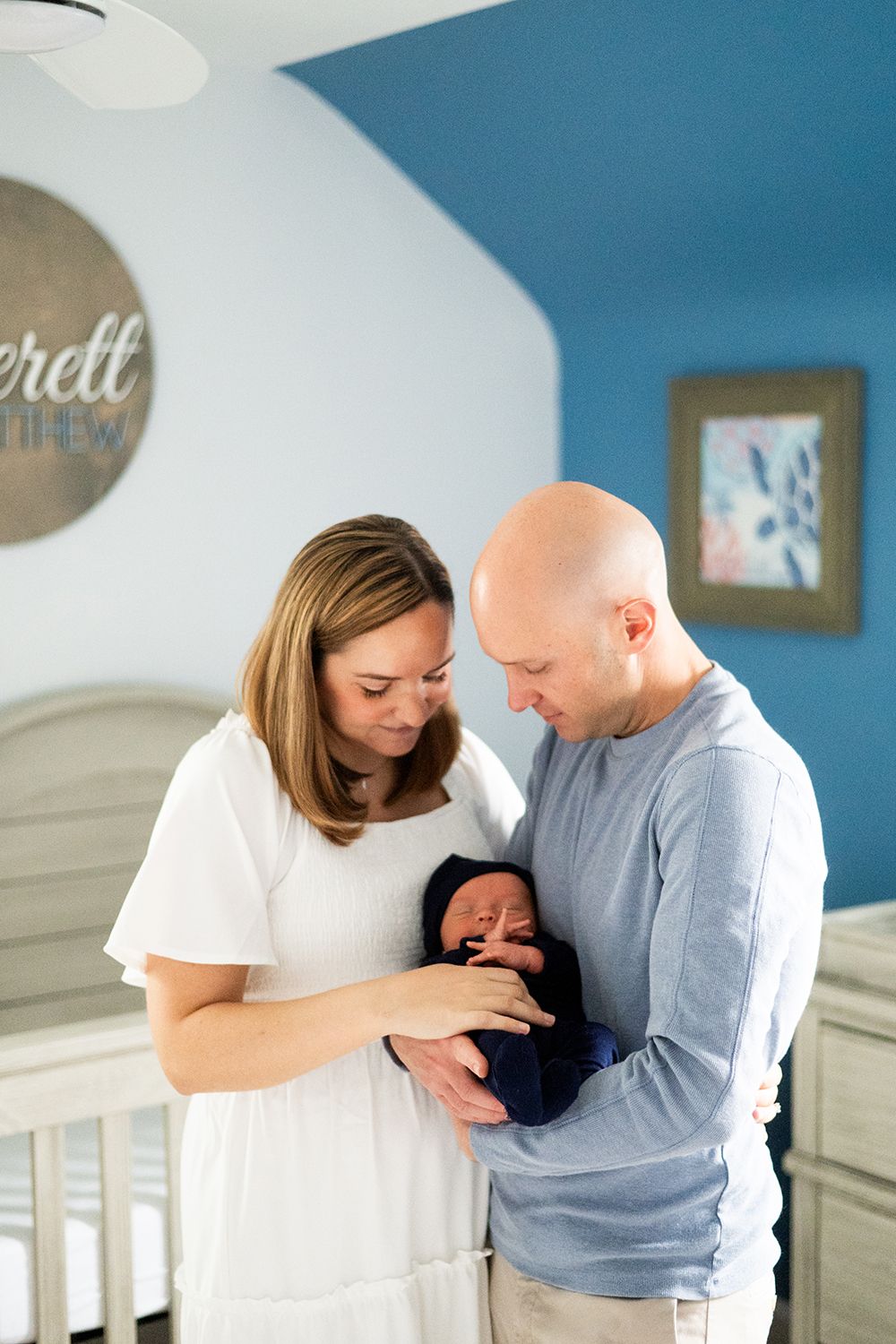Parents stand together in a nursery with blue walls, looking down lovingly at their newborn baby in a white crib.