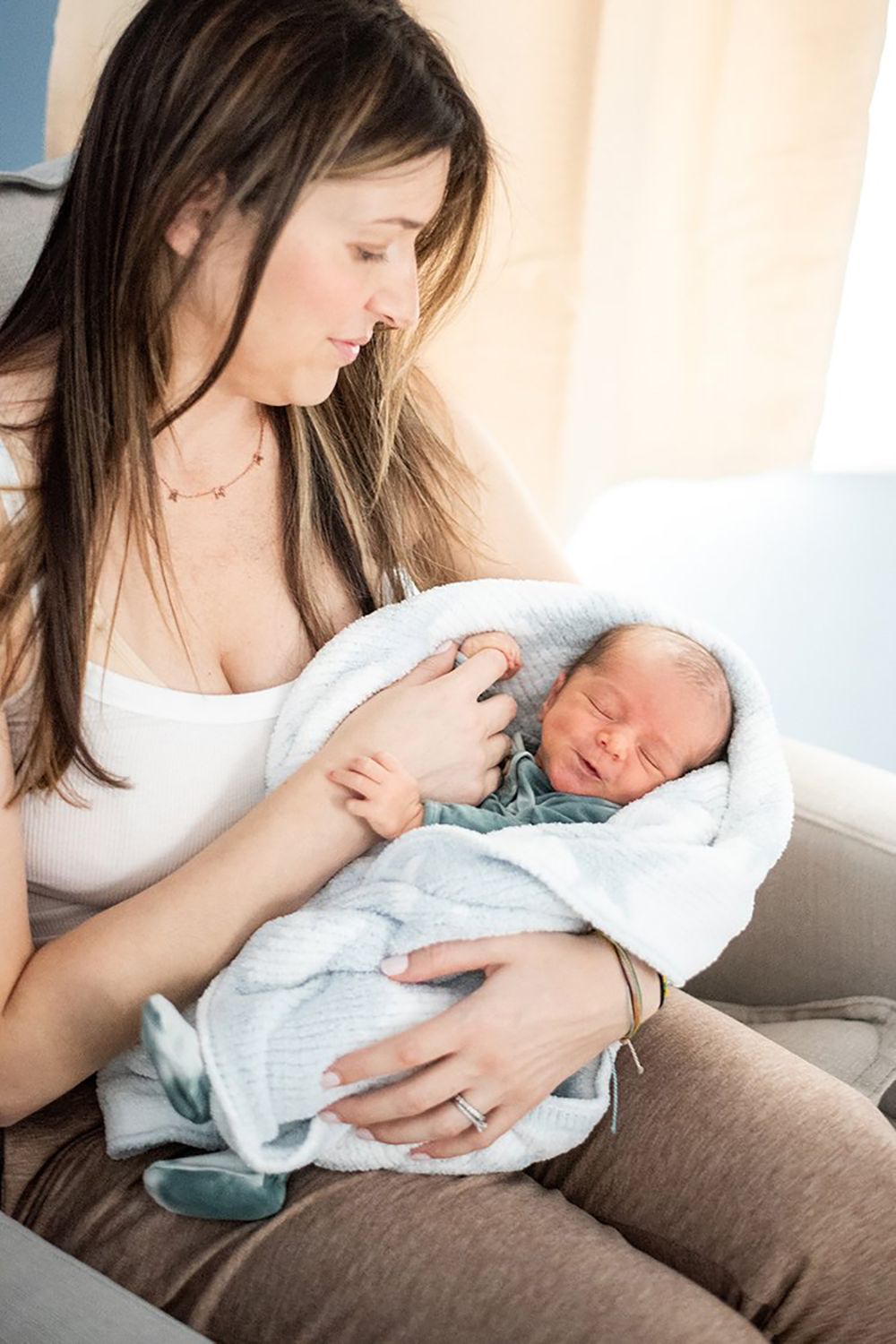 A mother in a white top tenderly holds and gazes at her sleeping newborn baby wrapped in a soft blanket.