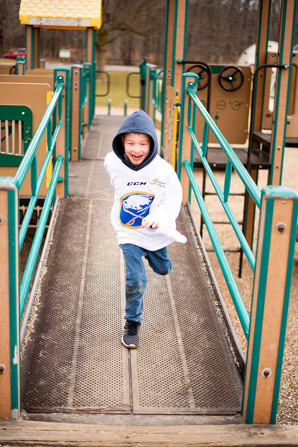 Young person in a white hoodie and jeans runs energetically across a playground ramp with green railings on a cloudy day.