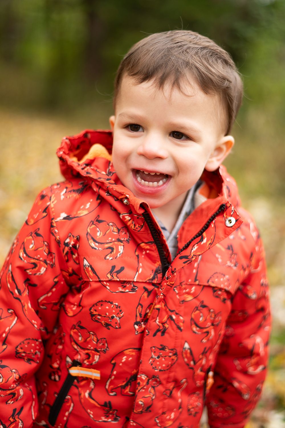 A cheerful toddler wearing a red patterned raincoat against a blurred autumn background.