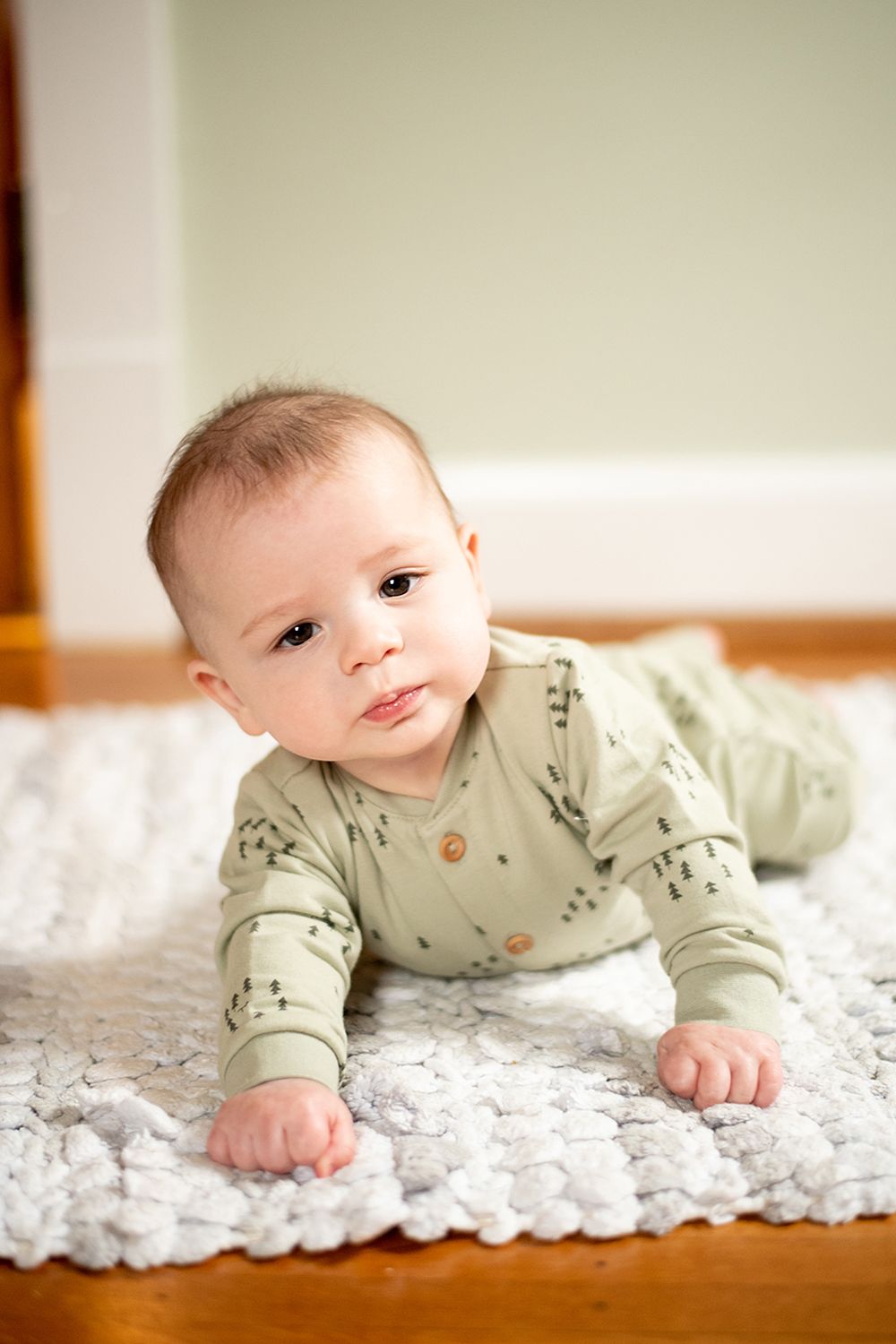 A young baby in sage green patterned sleeper outfit lies on their tummy on a white textured rug.