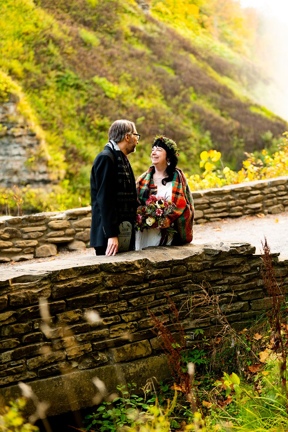 A romantic couple shares a moment on a stone wall surrounded by autumn foliage and golden sunlight.