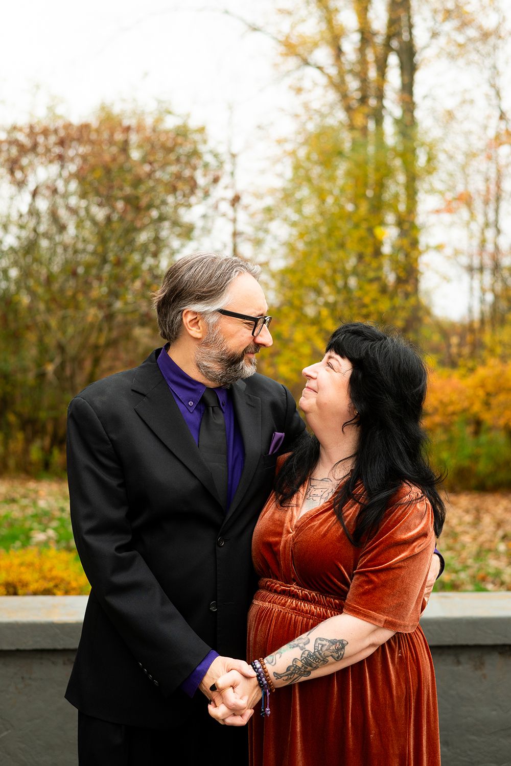 A couple in formal attire embrace outdoors in autumn, with one wearing black and purple and the other in a rust velvet dress.