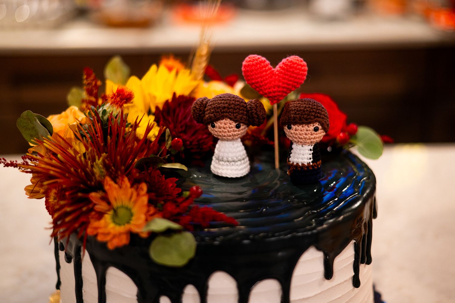 Black drip cake decorated with fall flowers and cute crocheted wedding cake toppers with a red heart accent.