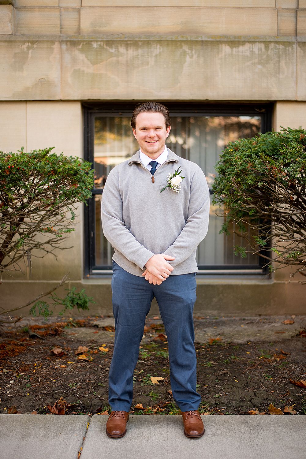 A person wearing grey sweater and navy pants poses in front of brick wall with cropped body parts shown.