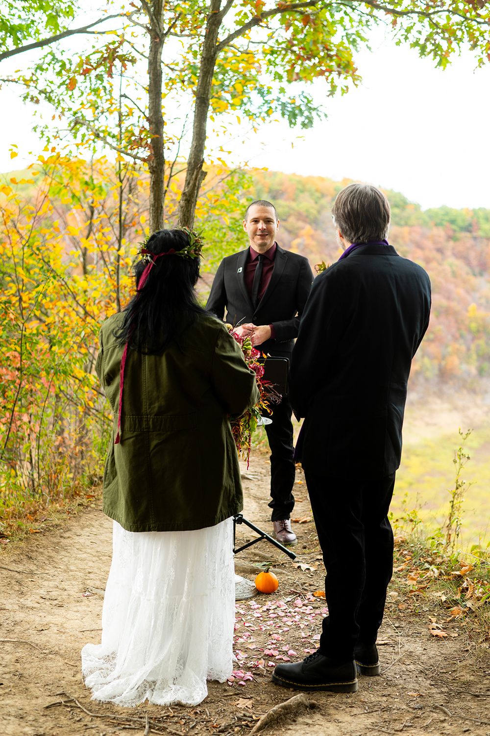 An outdoor autumn wedding ceremony takes place on a forest path with fall foliage in the background.