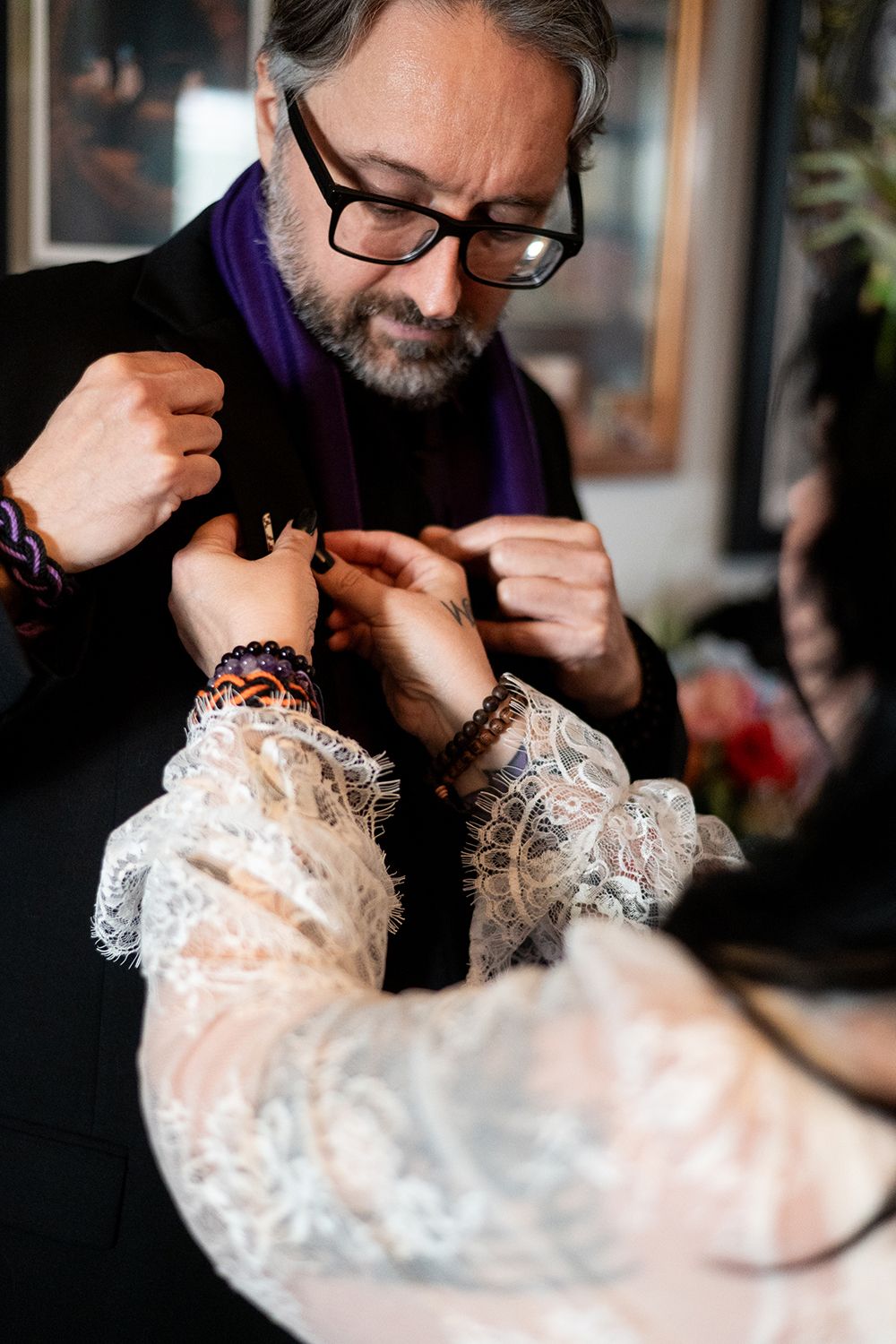 Person wearing glasses and dark coat fastens delicate lace dress sleeves during what appears to be a formal preparation.
