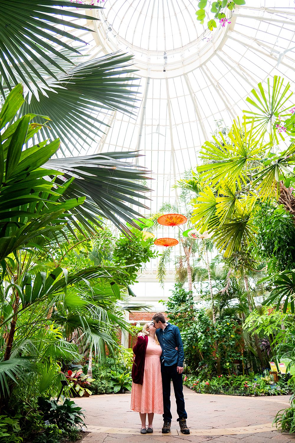 Couple shares romantic moment surrounded by lush palm fronds and tropical greenery in a botanical garden setting.