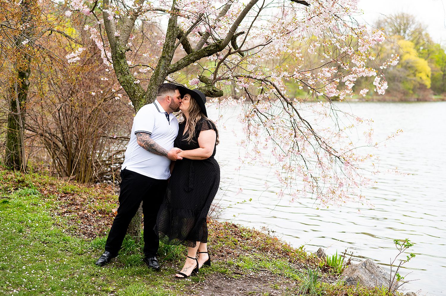 A couple embraces by a lake on a grassy shore during autumn with trees in the background.
