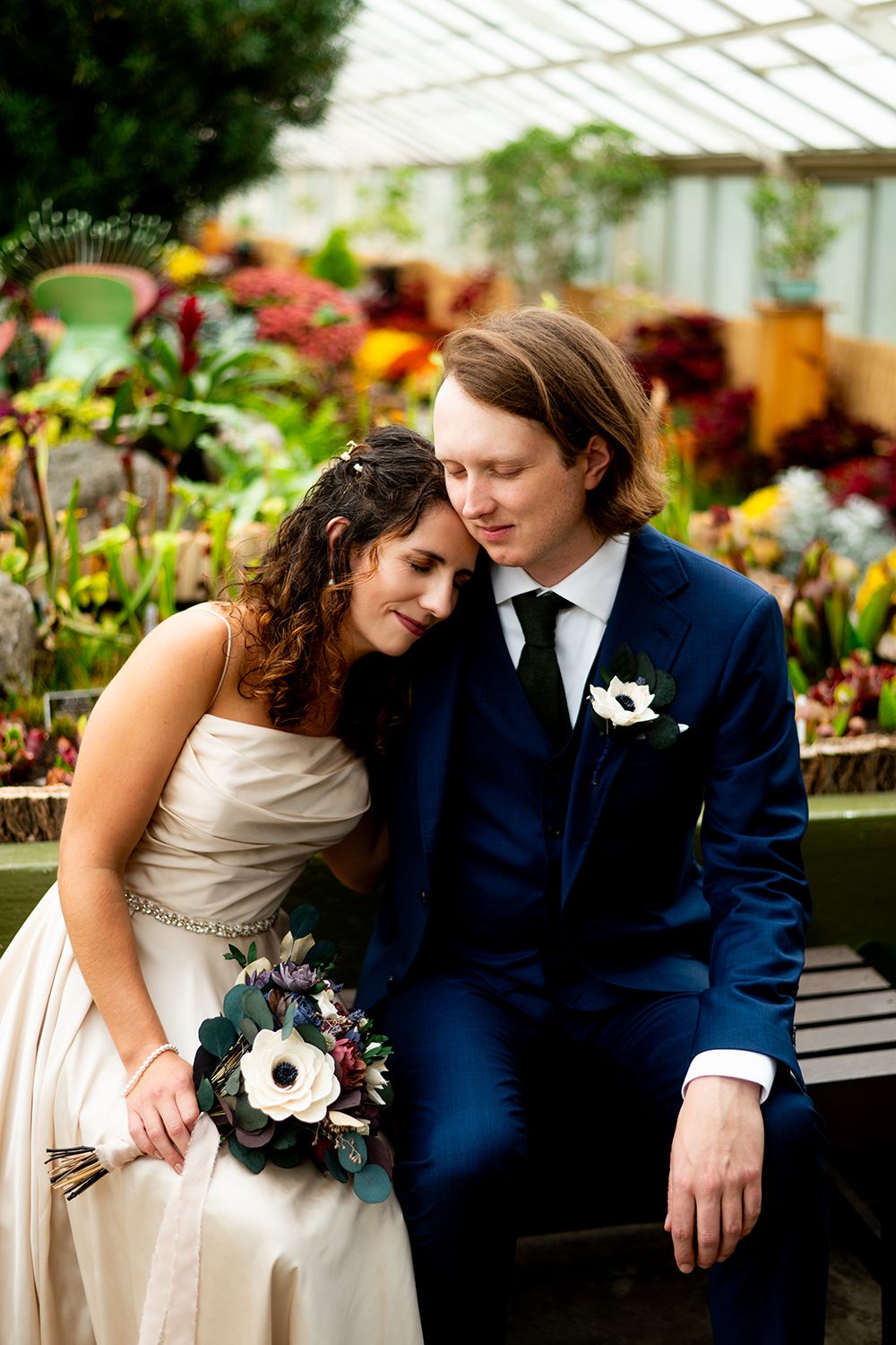 A romantic greenhouse wedding portrait with colorful flowers surrounding a couple in navy and cream attire.