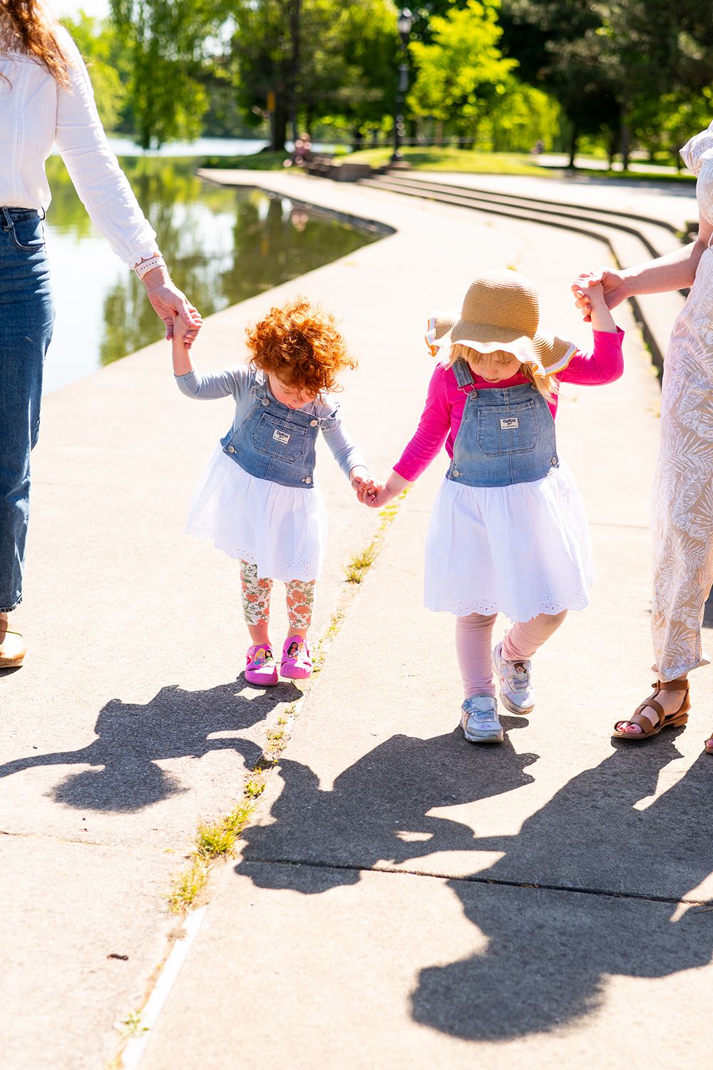 Two toddlers in matching denim outfits holding hands while walking with adults on a sunny path by a pond.