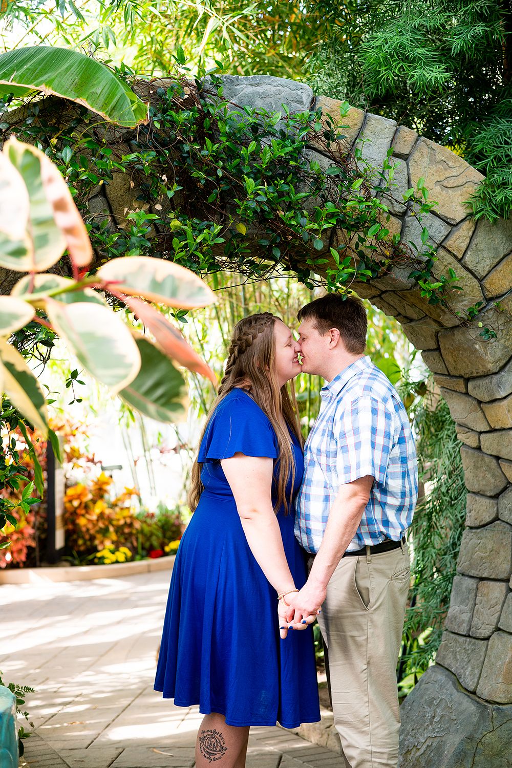 A couple shares a romantic kiss under a stone archway in a lush garden setting.