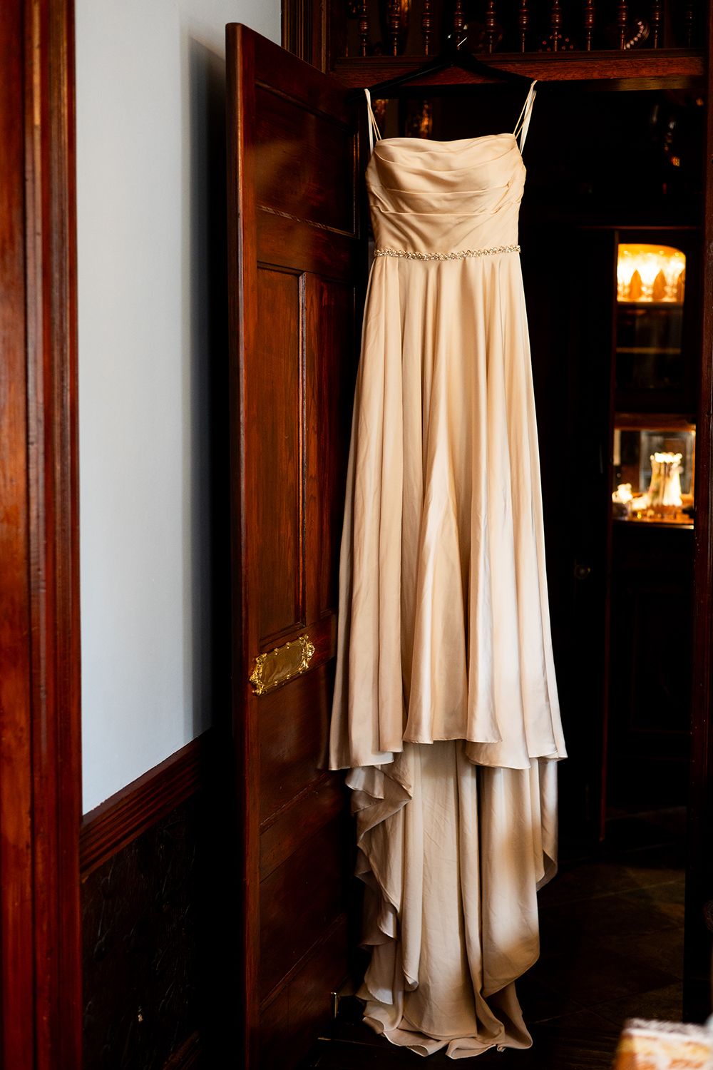 A beige chiffon bridesmaid dress hangs elegantly against a dark wooden wall in warm lighting.