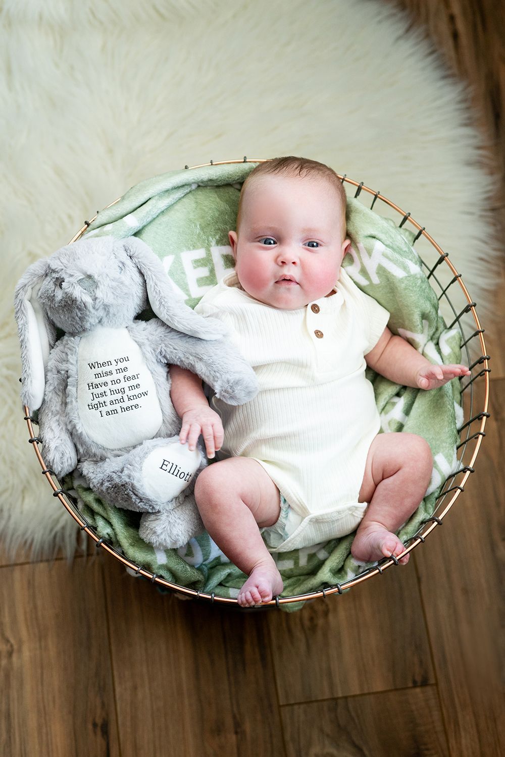 Baby in white onesie resting in round basket with plush bunny toy on furry white rug.