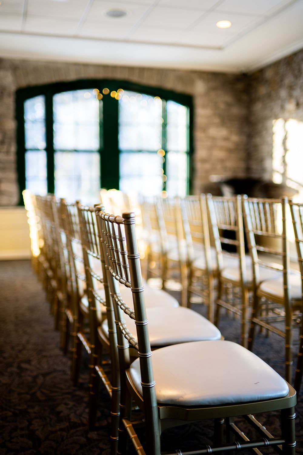 Row of gold Chiavari chairs with white cushions in an elegant indoor venue with exposed brick walls.