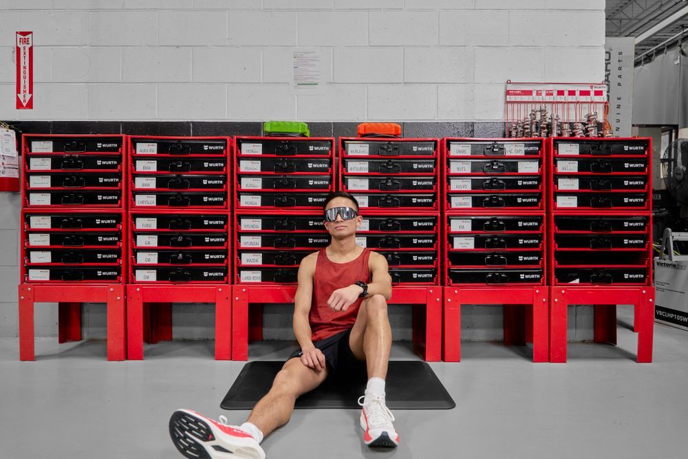 A man sits on the floor, leaning against a red storage unit, with a relaxed expression and casual clothing.