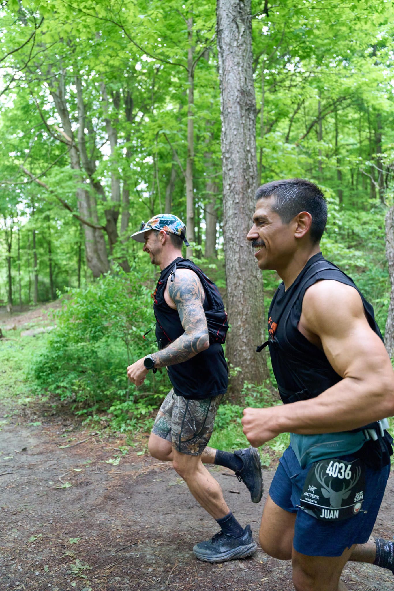 Two men running shirtless through a wooded area, surrounded by trees and natural scenery.