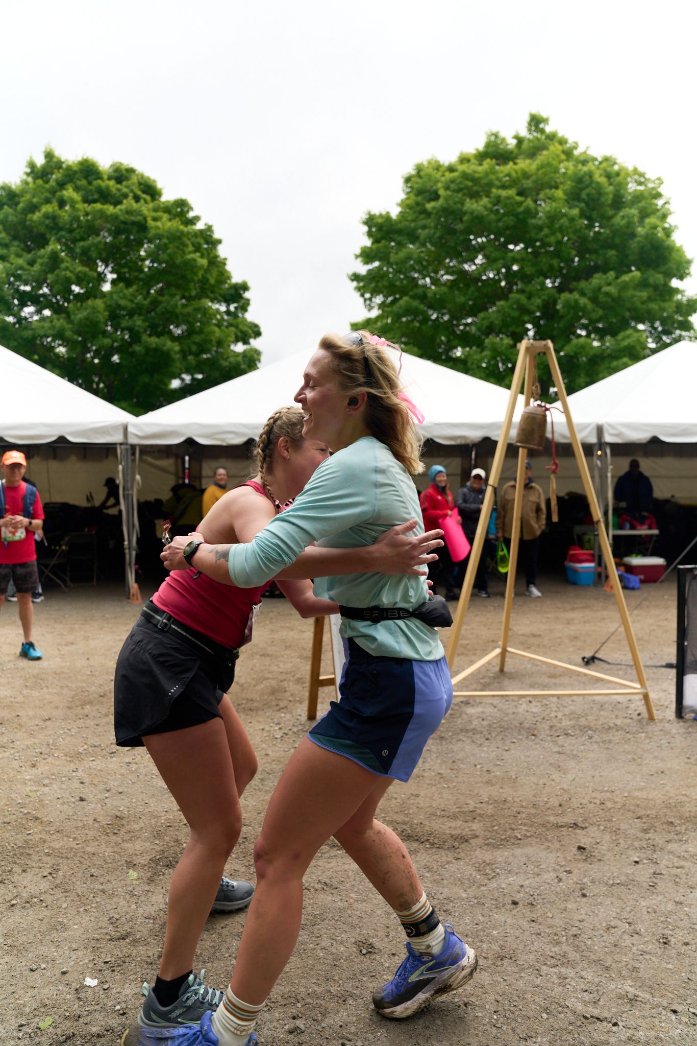 Two woman runs along a road, wearing a backpack, with trees lining the path in the background. Transition foa a relay race.