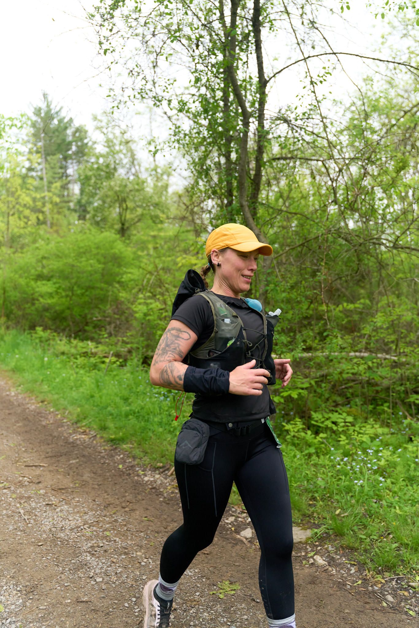 A woman runs along a road, wearing a backpack, with trees lining the path in the background.