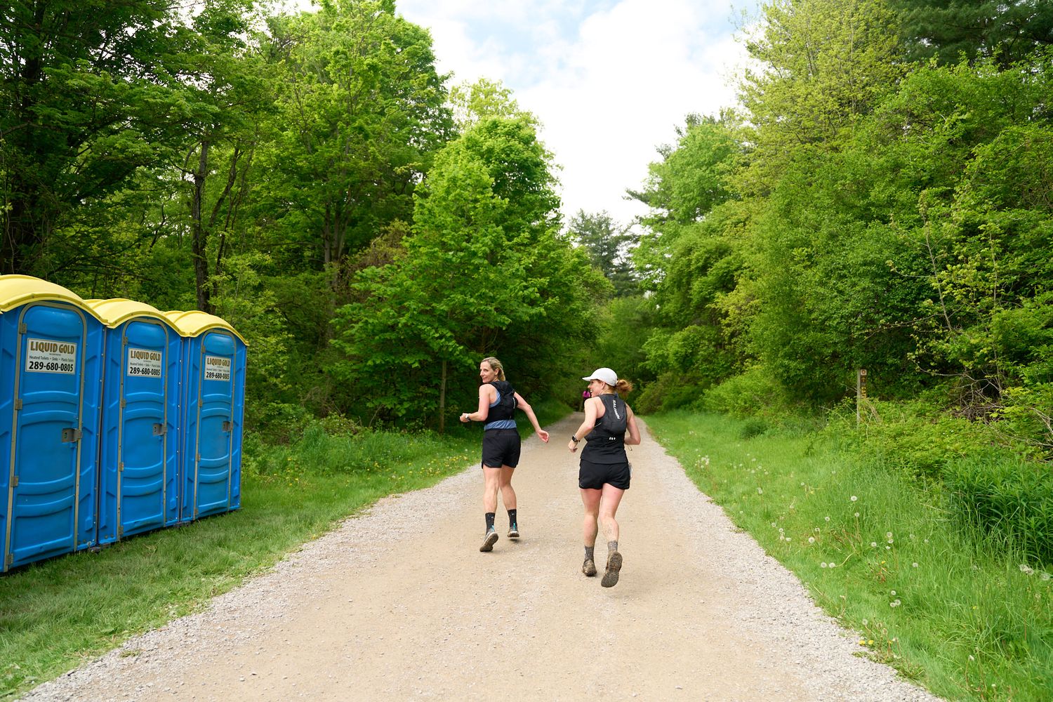 Two people racing on a dirt road adjacent to portable toilets, surrounded by greenery.