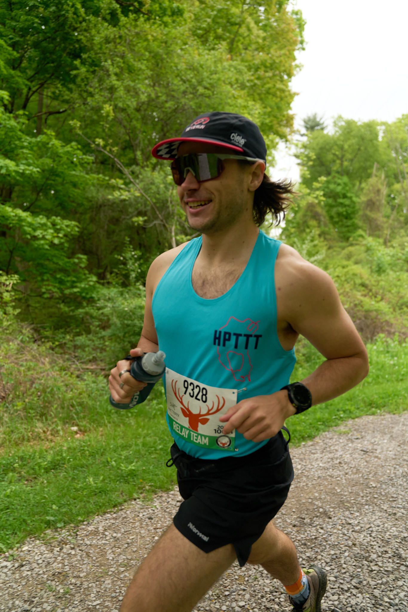 A man wearing a blue tank top and sunglasses is running outdoors.