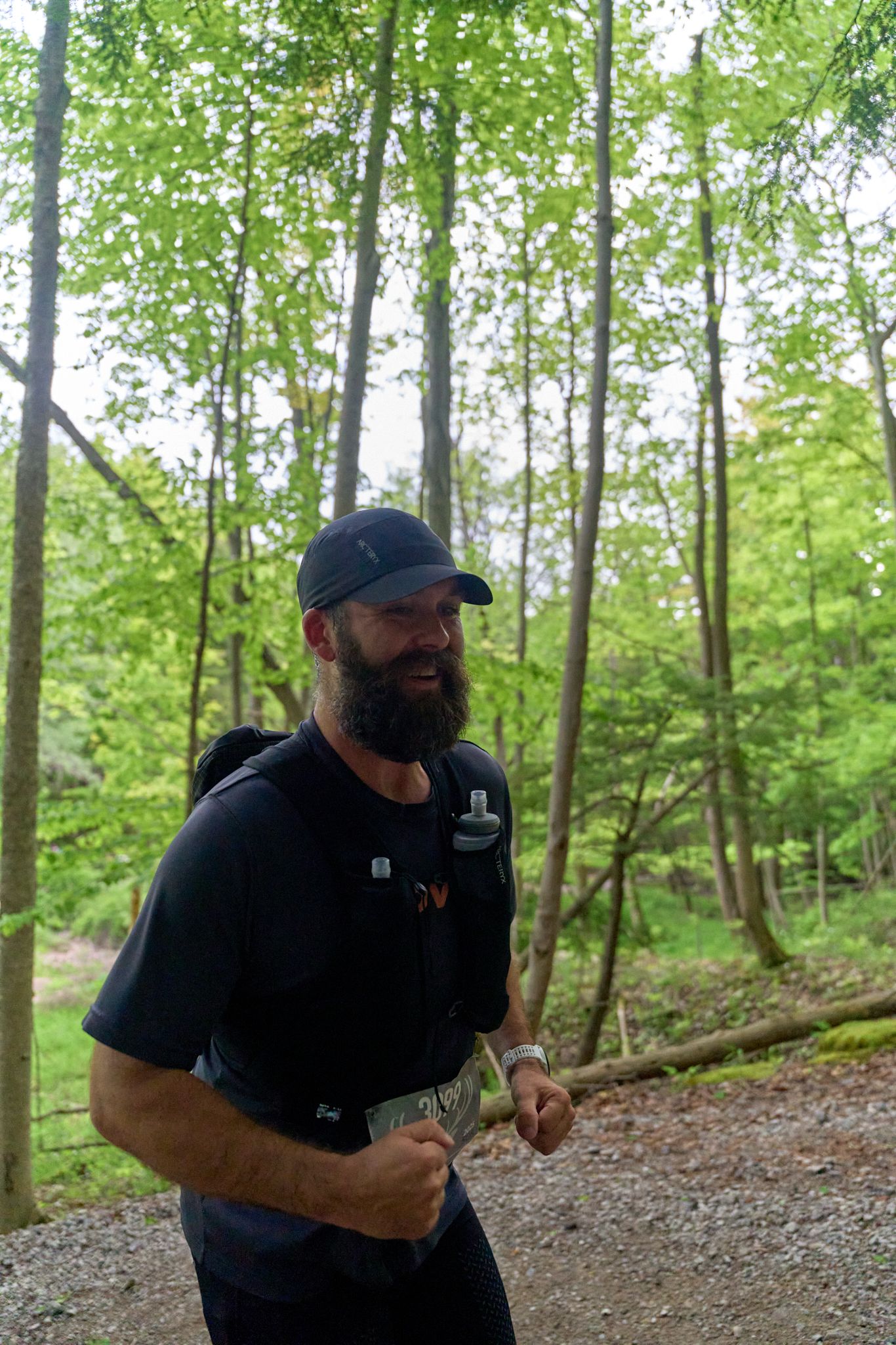 A bearded man in a backpack runs through a dense forest, surrounded by tall trees and dappled sunlight.