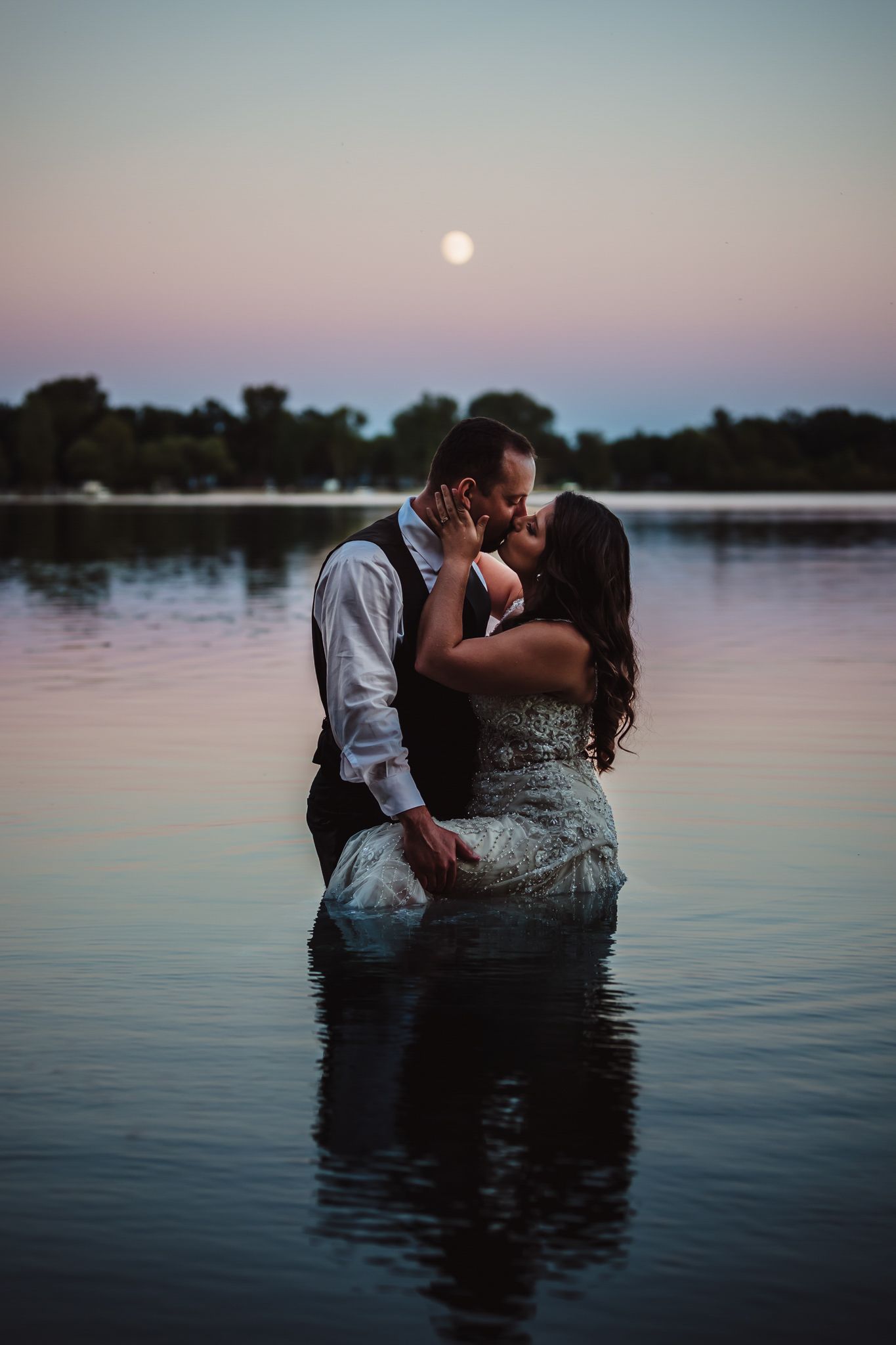 A romantic couple shares a kiss while standing in a lake at sunset with a full moon rising in the pastel sky.