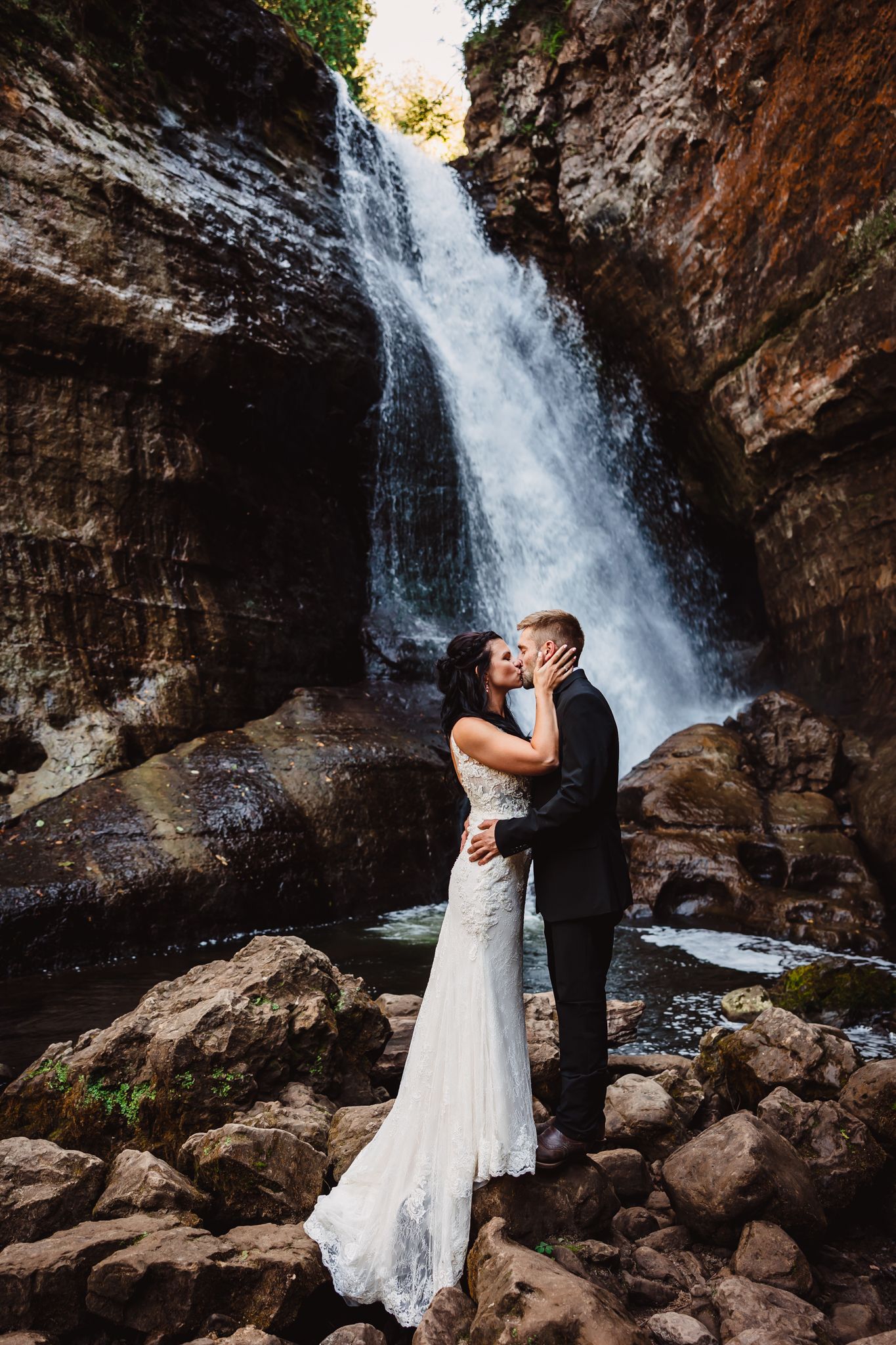 Couple embraces in front of a majestic waterfall cascading down rocky cliffs, surrounded by nature.