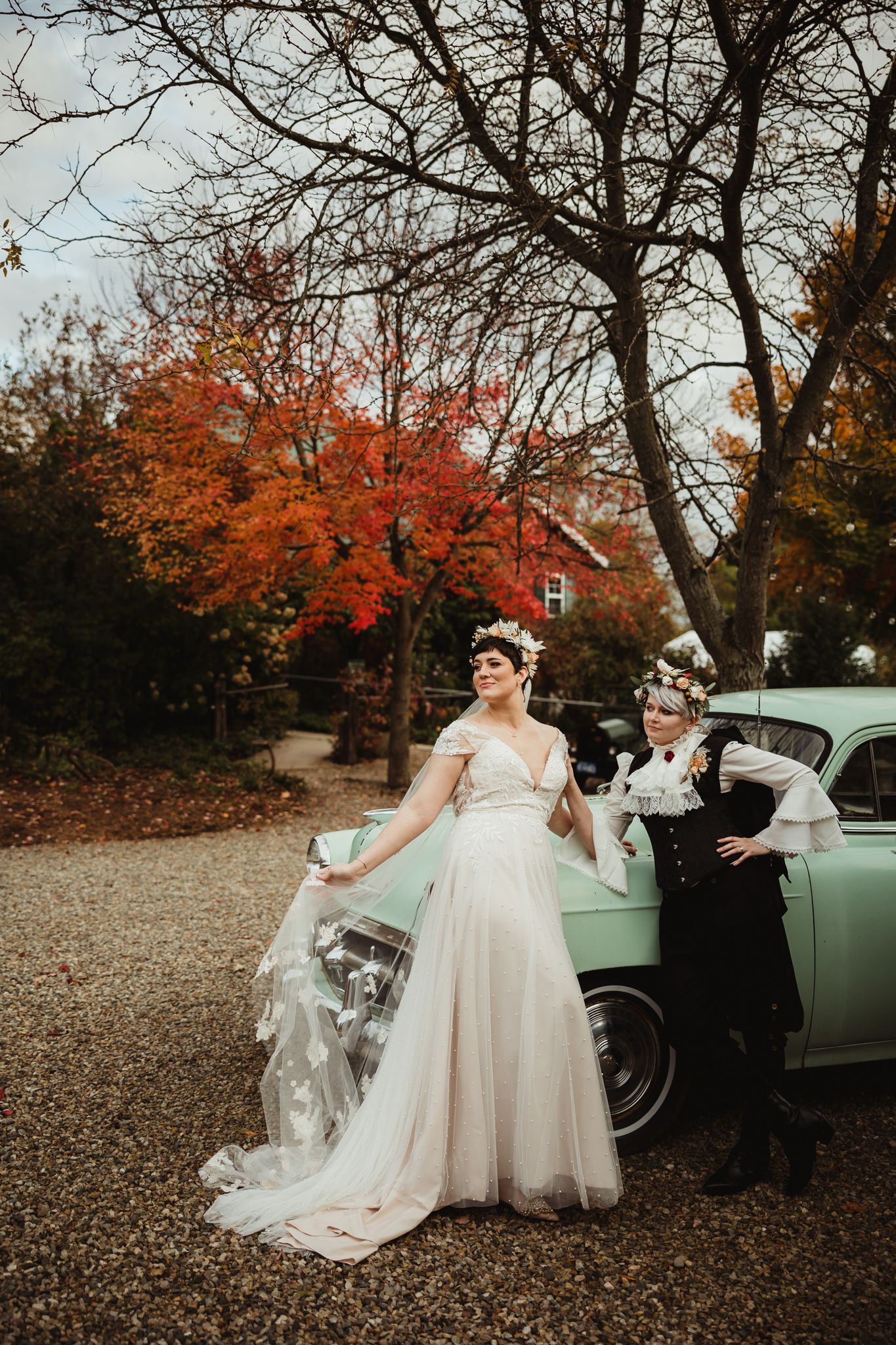 Two people pose with a vintage mint green car surrounded by autumn trees with vibrant red foliage.