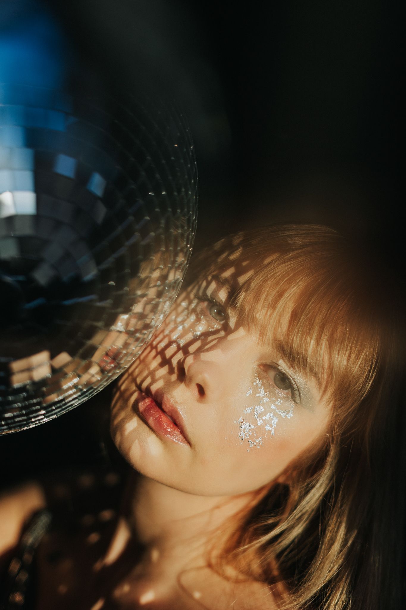 Glittery disco ball casts sparkling light patterns across person's face in dramatic nightclub setting.