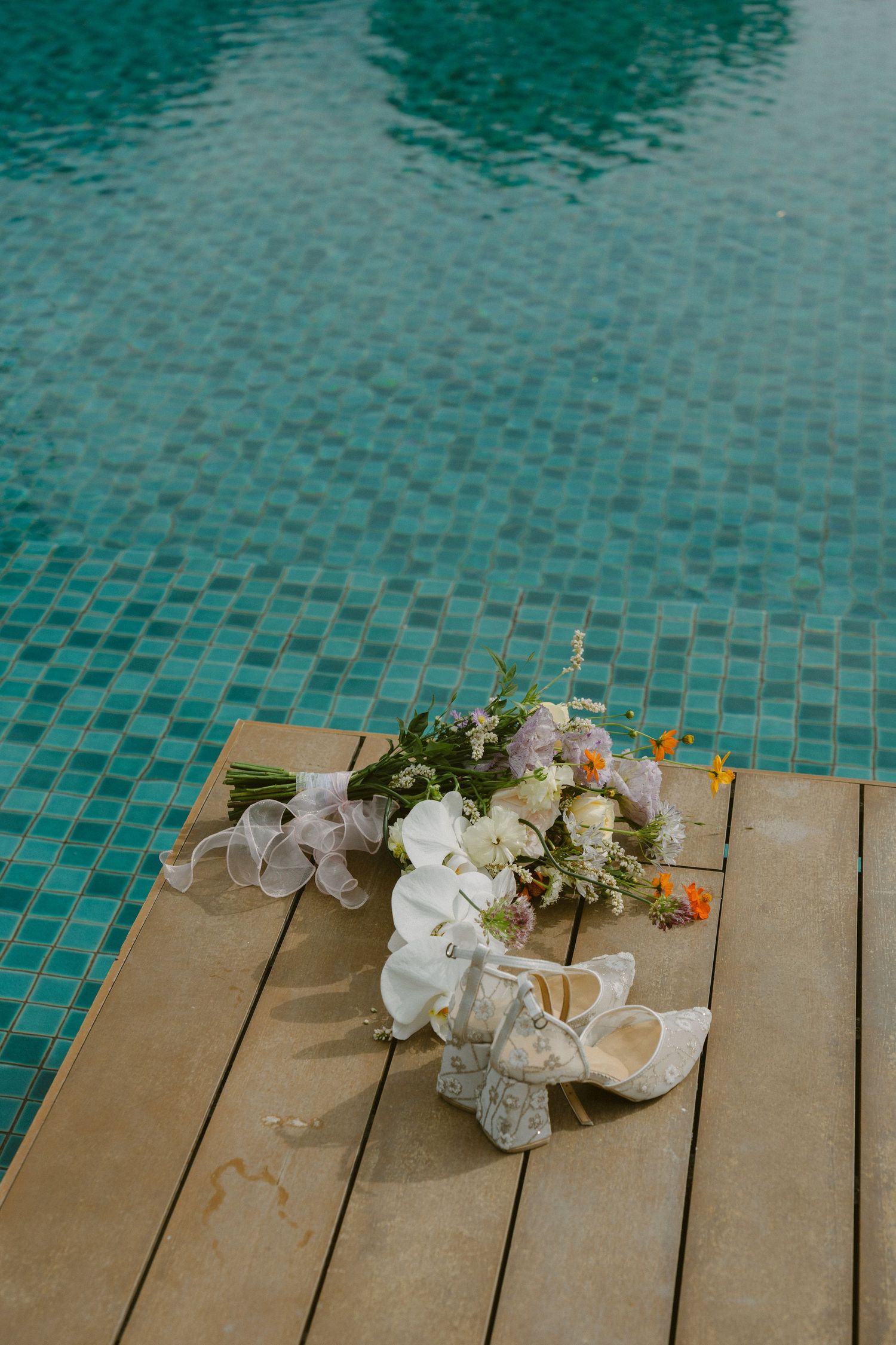 Wedding decor and empty glasses scattered on wooden decking beside turquoise pool water.