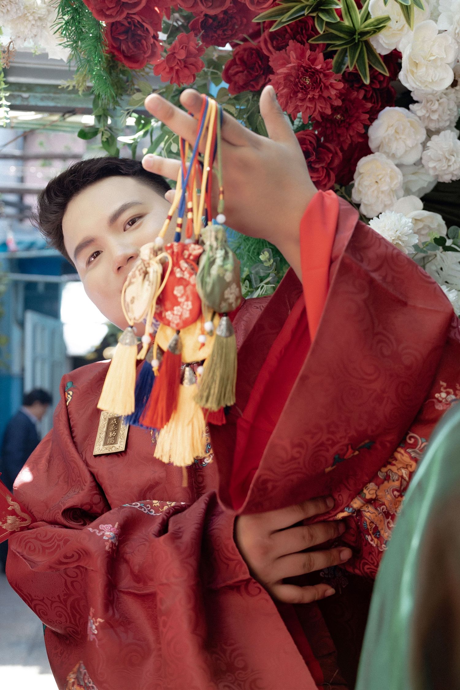 A person in a red traditional Chinese outfit holds decorative tassels against a floral backdrop with red and white flowers.