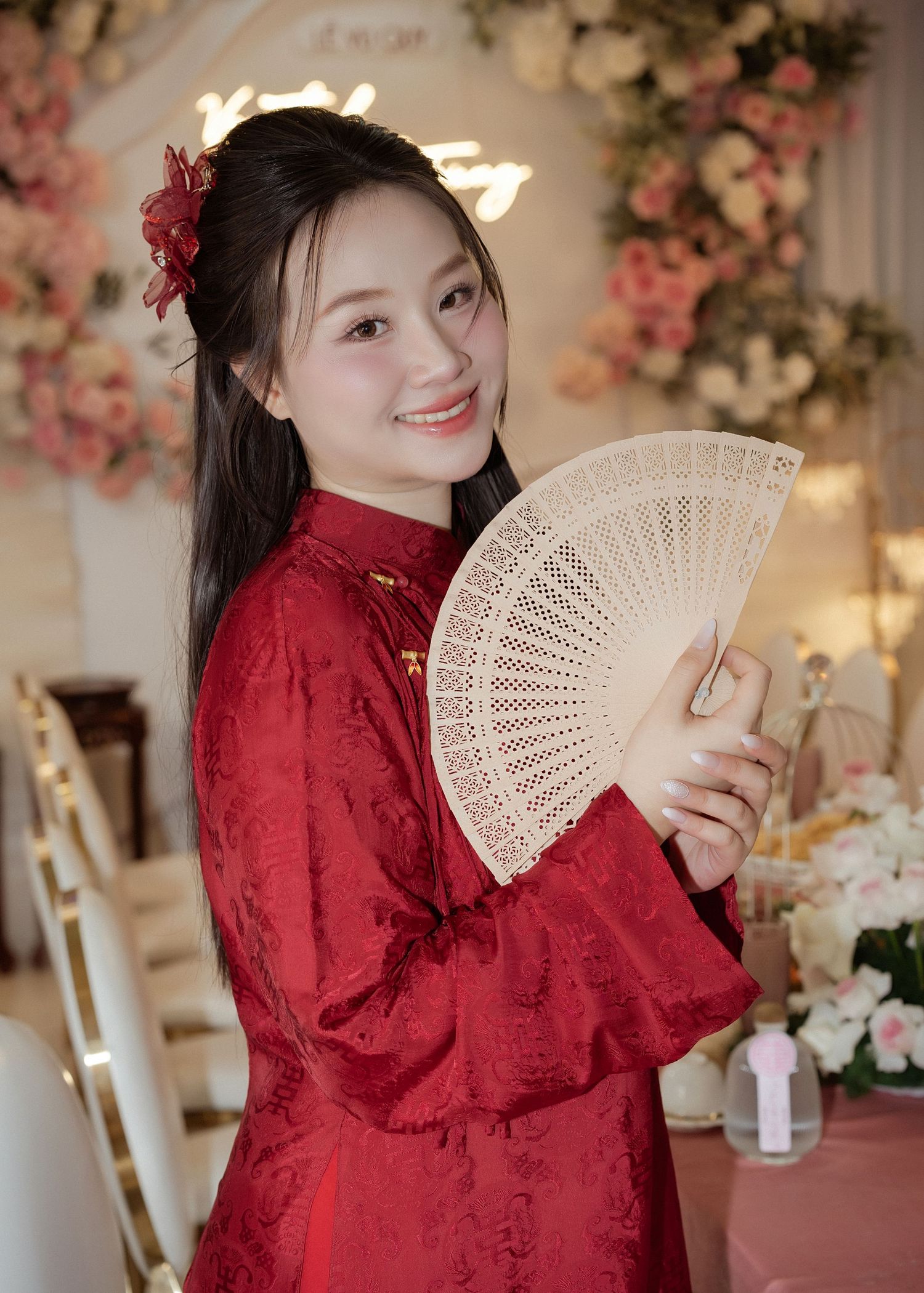 Person in traditional red Chinese qipao dress holding an ornate white folding fan against floral backdrop.