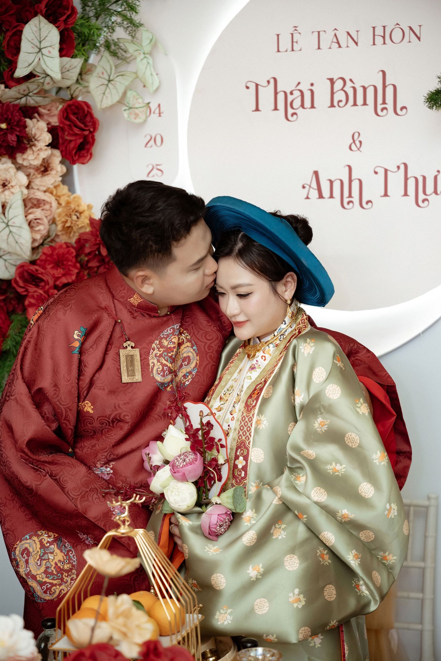 A couple in traditional Vietnamese wedding attire share an intimate moment against a floral backdrop with red and pink roses.