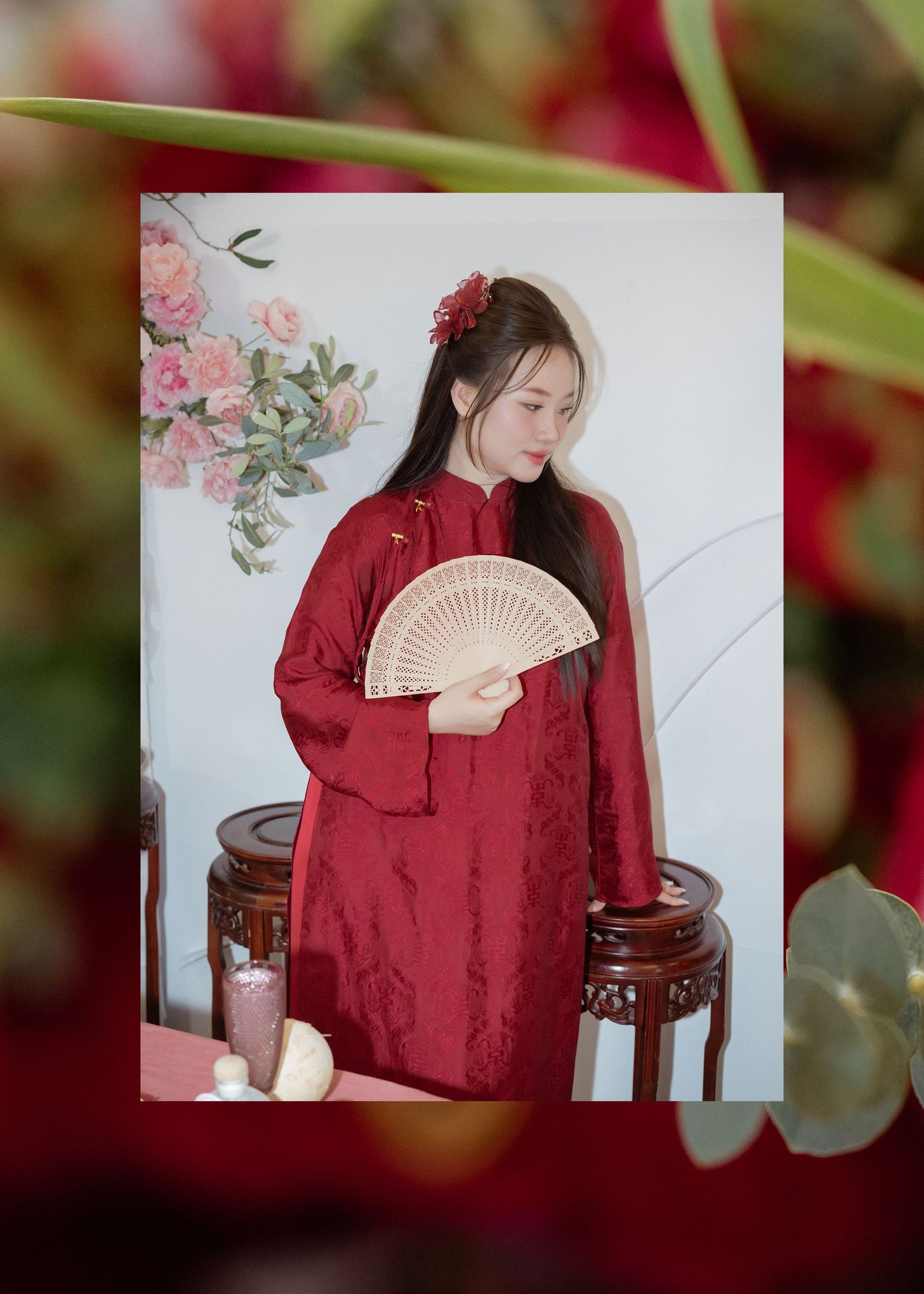 A figure in traditional Asian style red hanfu dress holds a folding fan while posing elegantly against a floral backdrop.