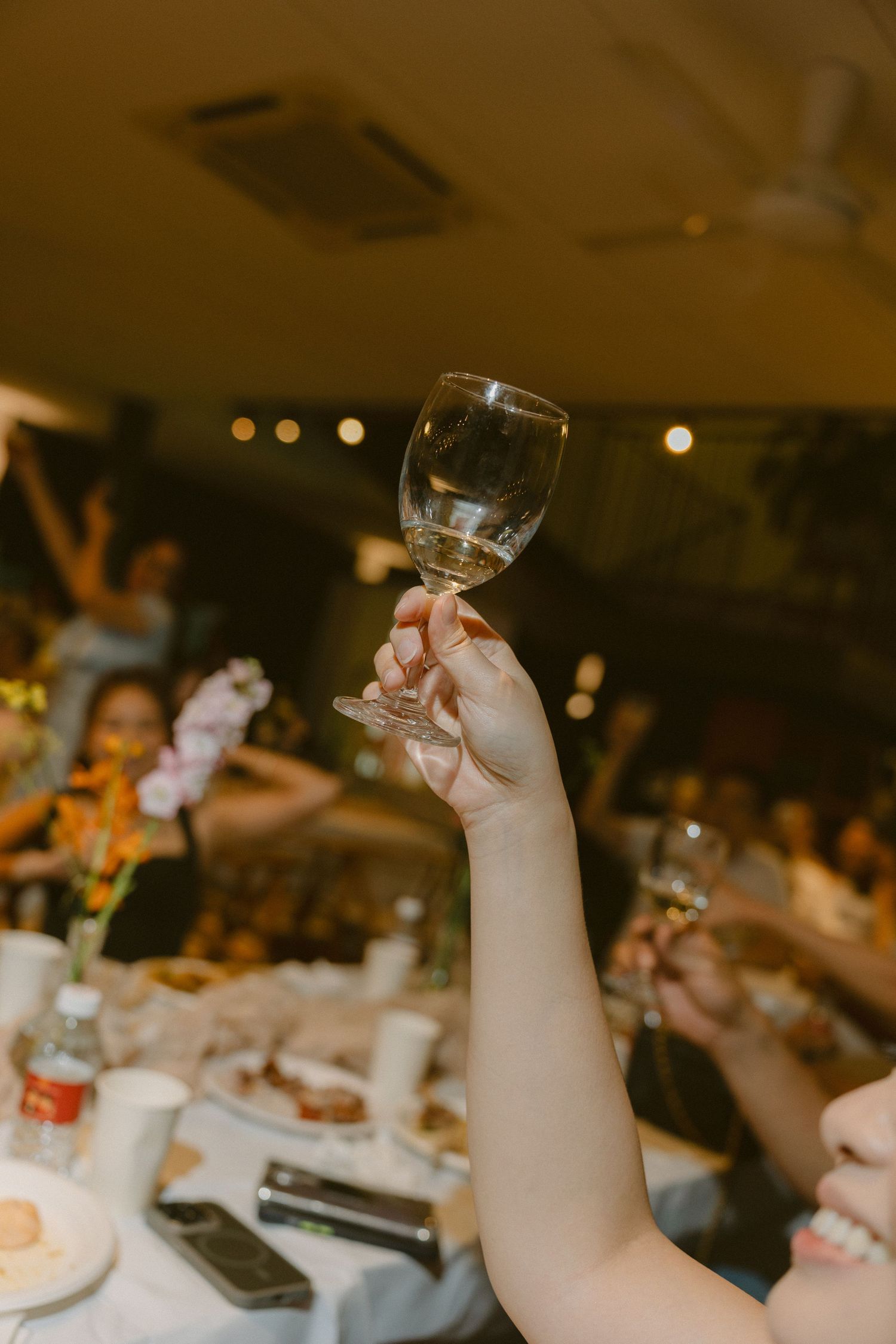 A hand raises a wine glass during a wedding reception toast with bokeh lights and table settings in the background.