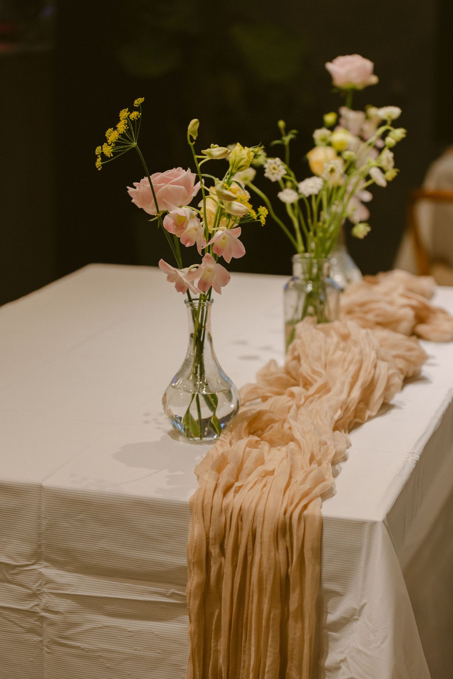 Romantic table setting with soft pink flowers in glass vases and flowing peach fabric runners on white tablecloths.