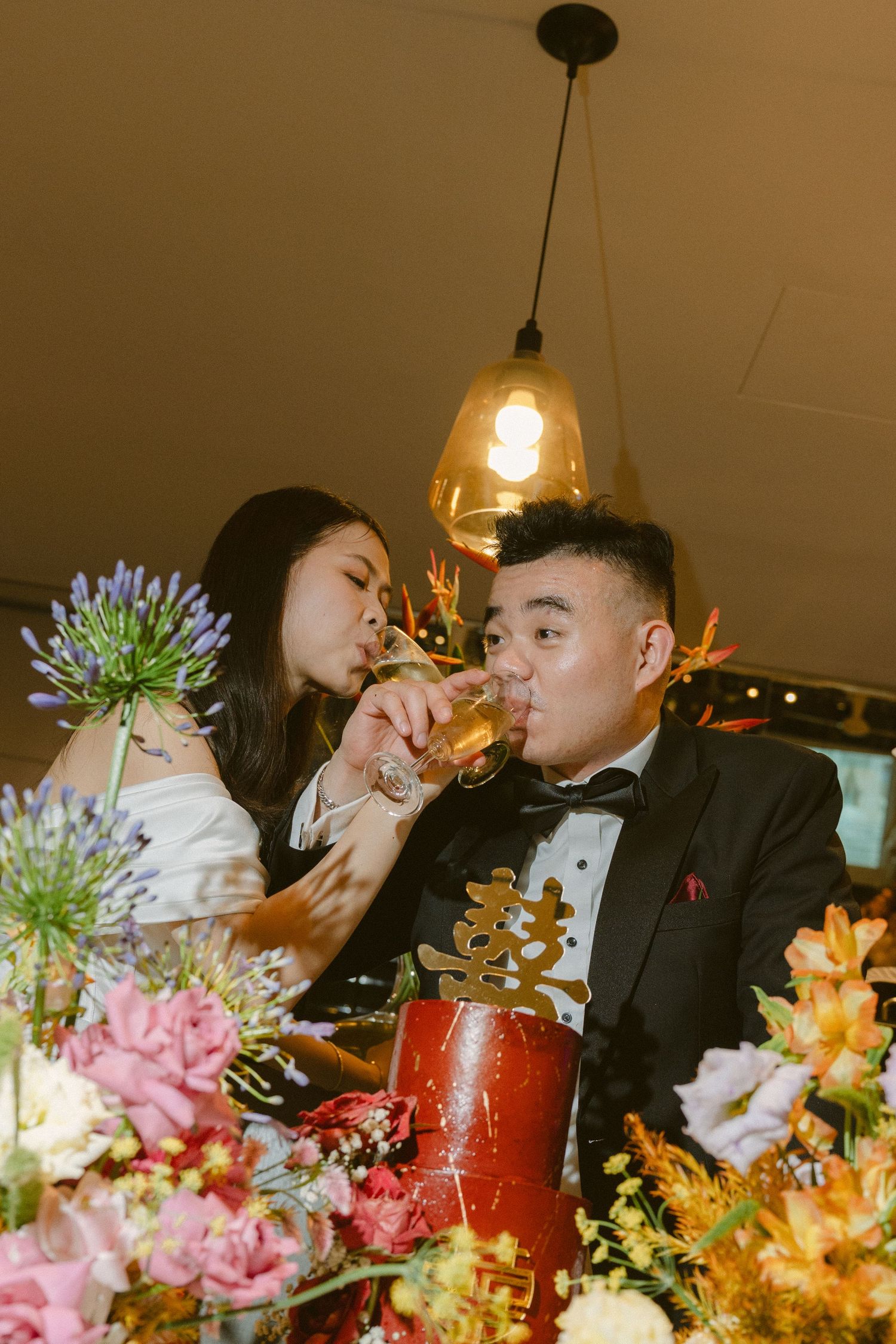 Wedding guests enjoy drinks at a reception table decorated with colorful floral arrangements and glowing pendant lights.